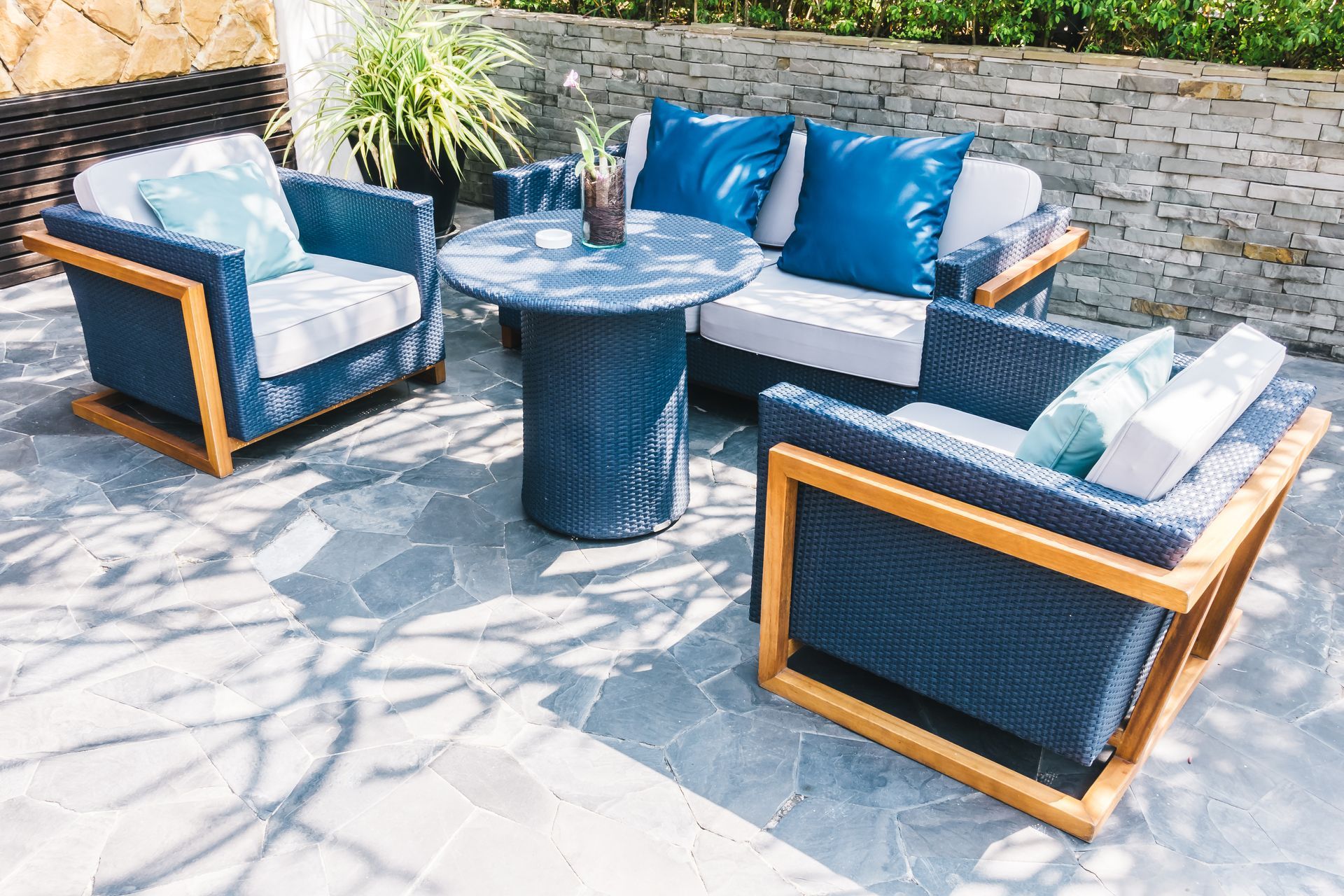 Outdoor patio seating set with blue-gray cushions, wooden chairs, and a round table against a stone wall.