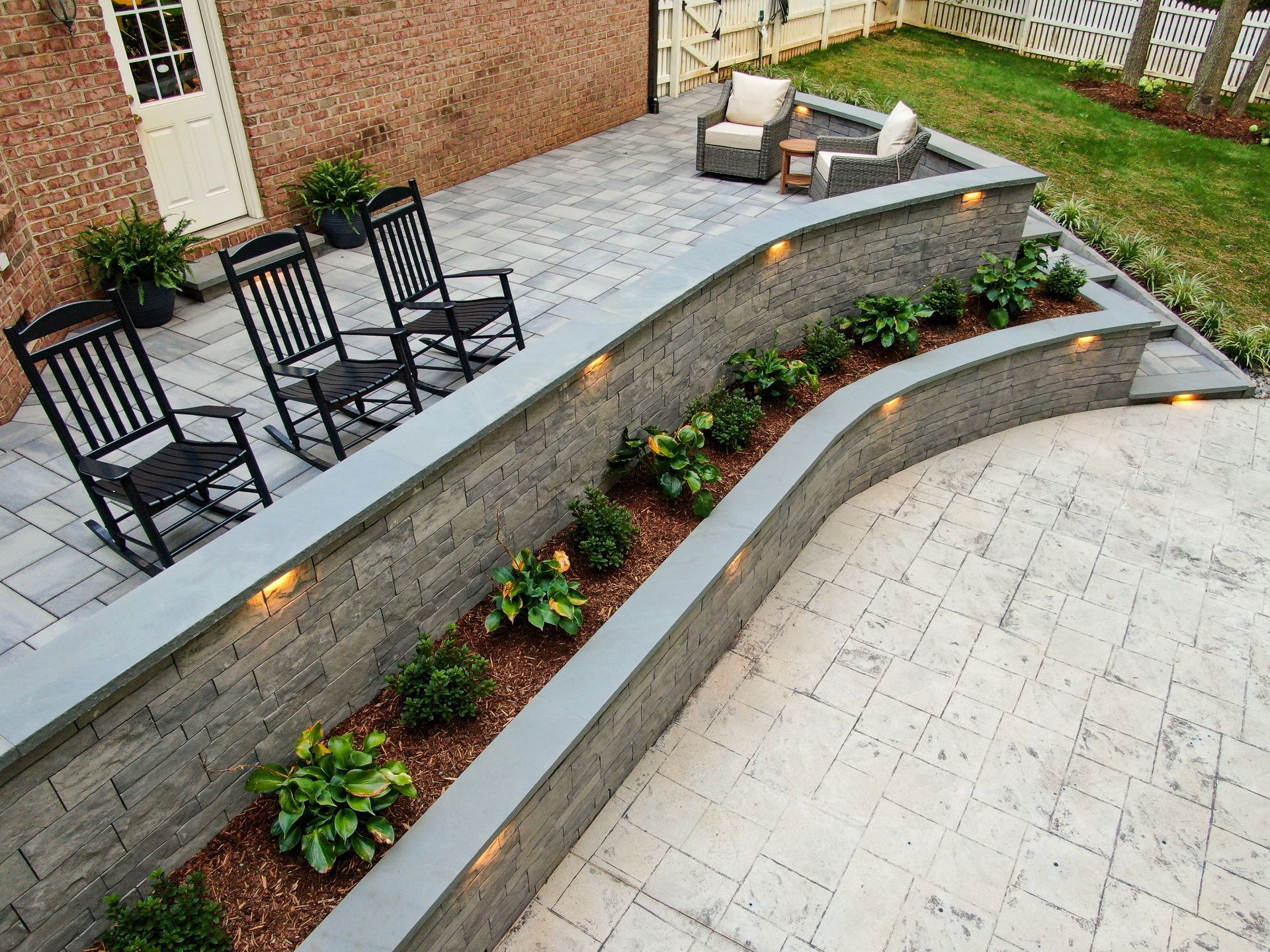 Curved stone patio with black chairs, raised planter beds, and a tiled courtyard below