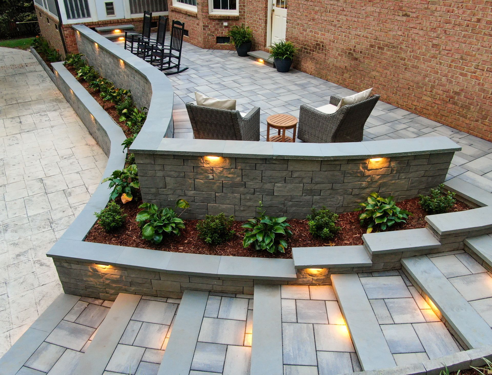 Outdoor stone patio with curved retaining wall, steps, built-in lights, and seating area with wicker chairs and table