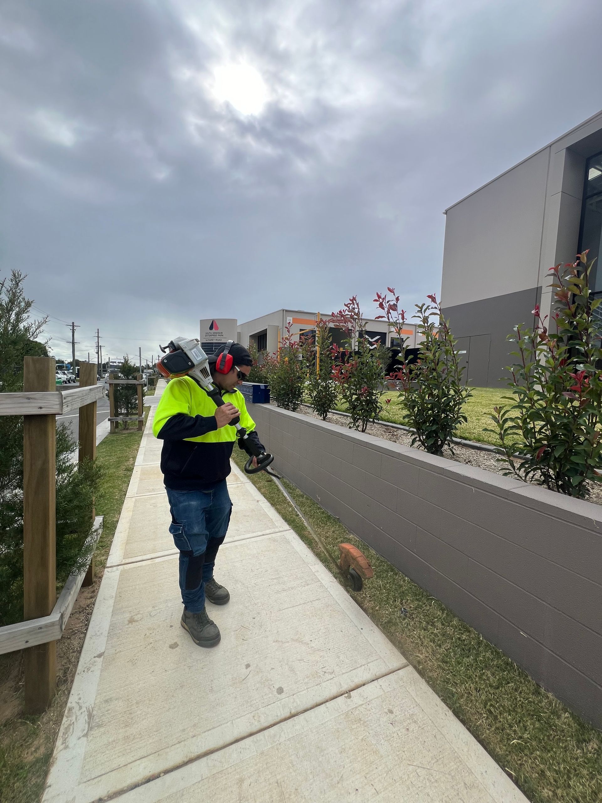 A man is standing on a sidewalk cutting grass with a lawn mower.