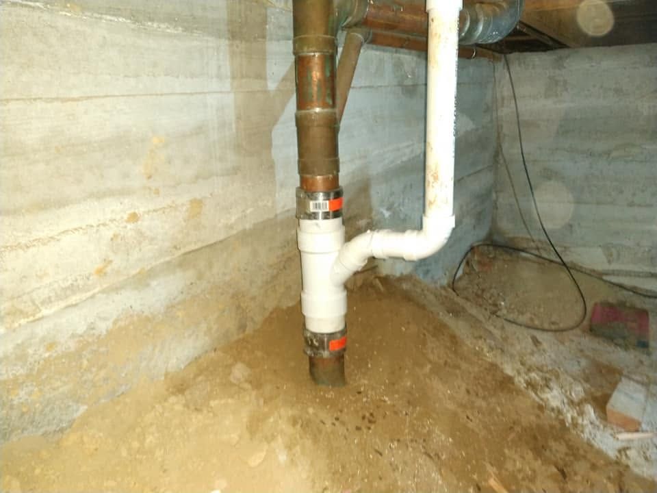 Underground utility space with copper and PVC plumbing pipes against a concrete wall, surrounded by dirt