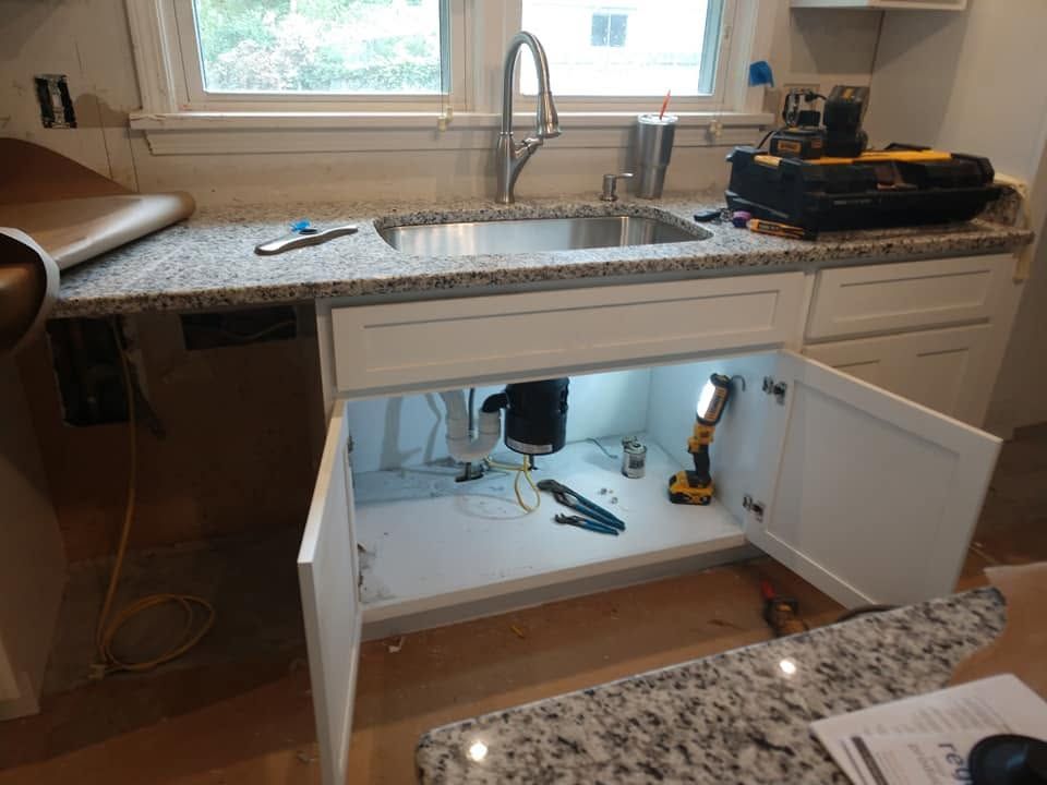 Kitchen sink cabinet under repair, with open doors exposing pipes and tools beneath a granite countertop.
