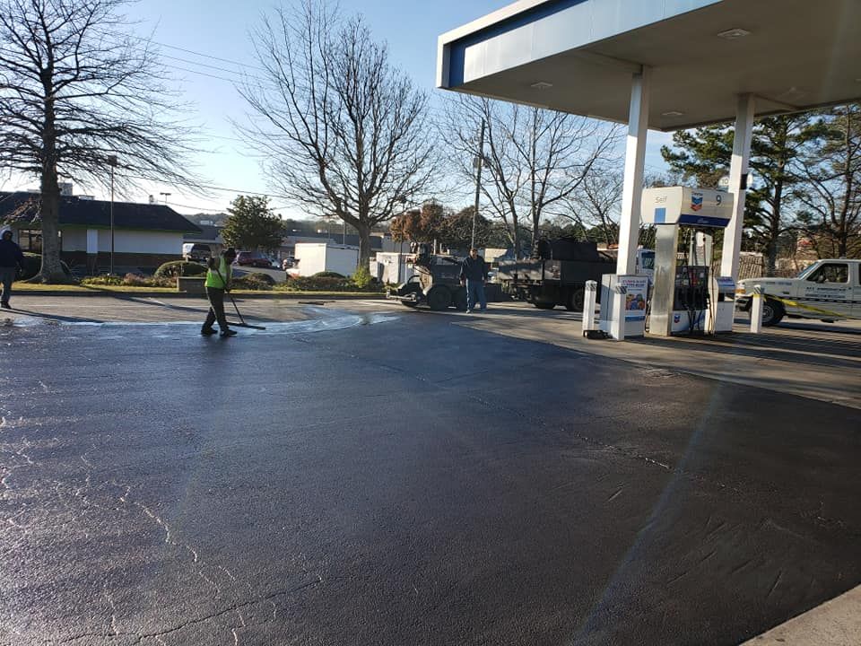 Gas station with workers cleaning wet pavement. Cars, trees, and blue sky visible.