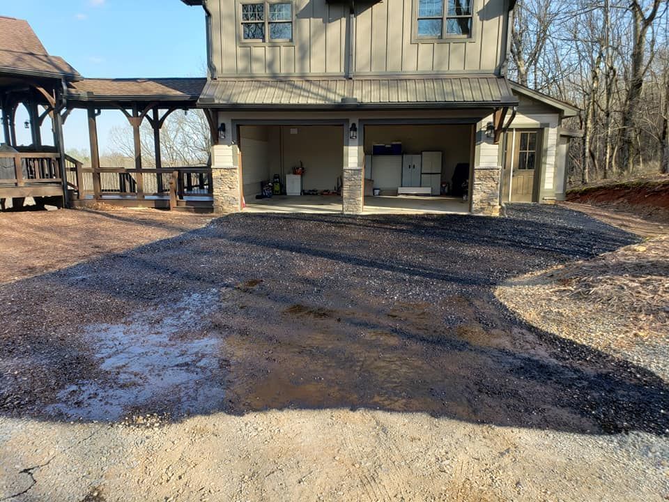 Asphalt driveway leading to a garage under a two-story house with a wooden porch on the left.