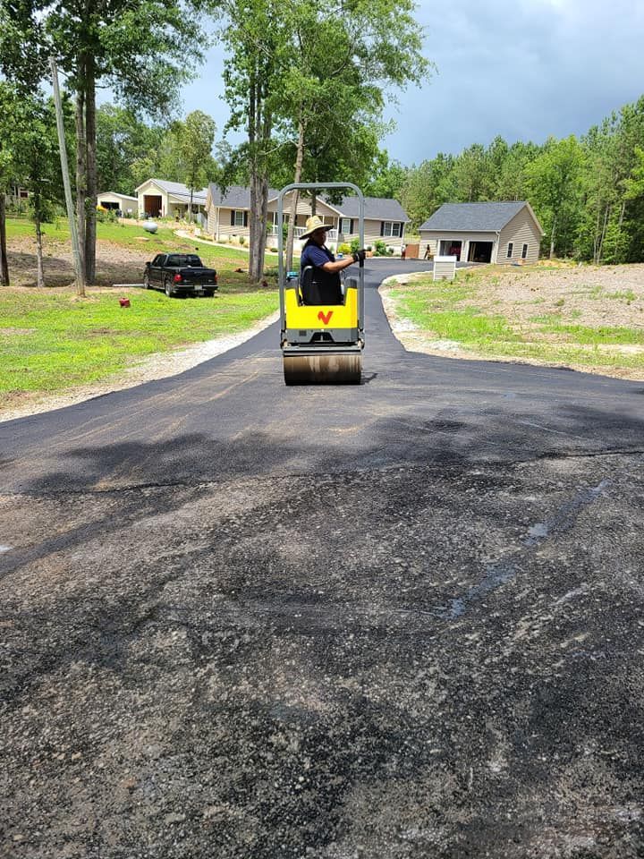 Person operating a road roller on freshly paved asphalt road; residential homes in the background.