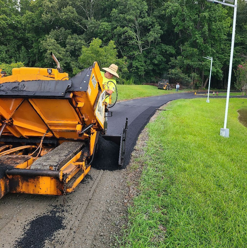 Yellow asphalt paving machine laying new asphalt on a path next to a green grassy area.