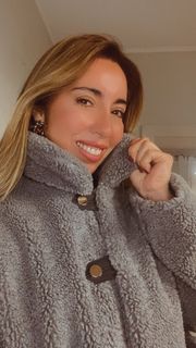 Persona sonriente con un abrigo gris de pelo sintético en el interior de un edificio, sosteniendo el cuello de la prenda hacia arriba.