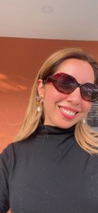Una persona sonriente con gafas de sol y un jersey de cuello alto negro, frente a una pared de color naranja cálido.