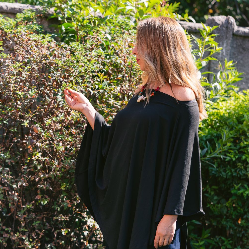 Mujer con vestido negro de pie en un jardín soleado, mirando un arbusto frondoso.