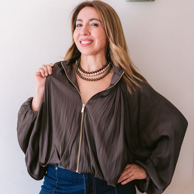 Mujer con blusa oscura y vaqueros azules, sonriendo con una mano en la cadera sobre un fondo claro.
