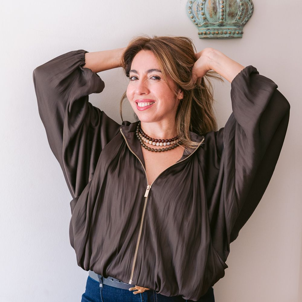 Mujer sonriente con blusa oscura y vaqueros, posando con las manos detrás de la cabeza contra una pared clara.