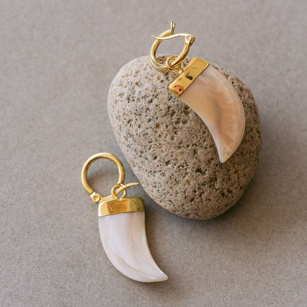 Pendientes de aro dorados con colgantes de concha blanca y melocotón sobre un fondo de piedra gris.