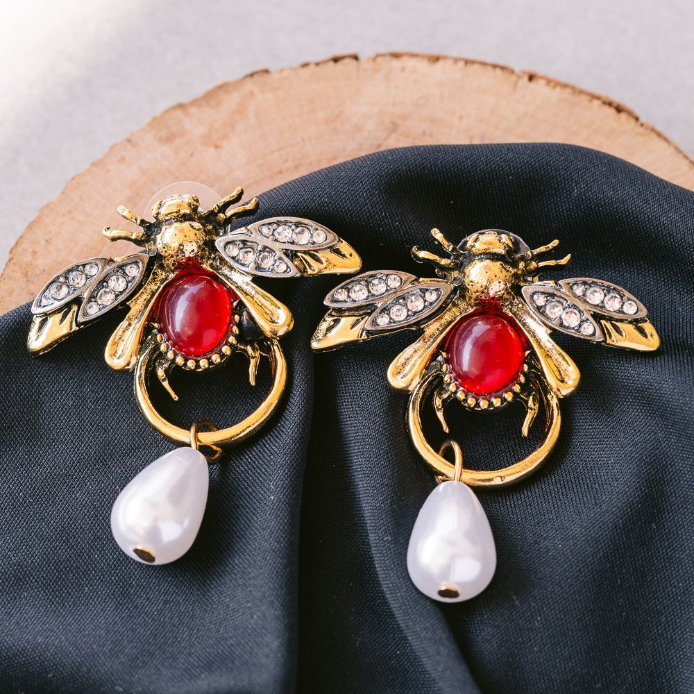 Par de pendientes con forma de abeja adornados con piedras rojas y perlas blancas sobre tela negra.
