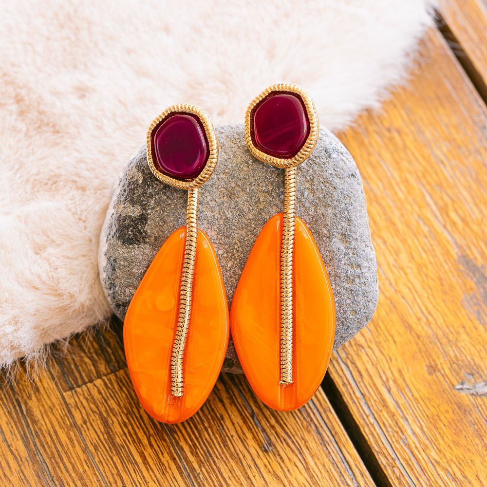 Pendientes colgantes naranjas y dorados con piedras burdeos sobre una piedra gris.