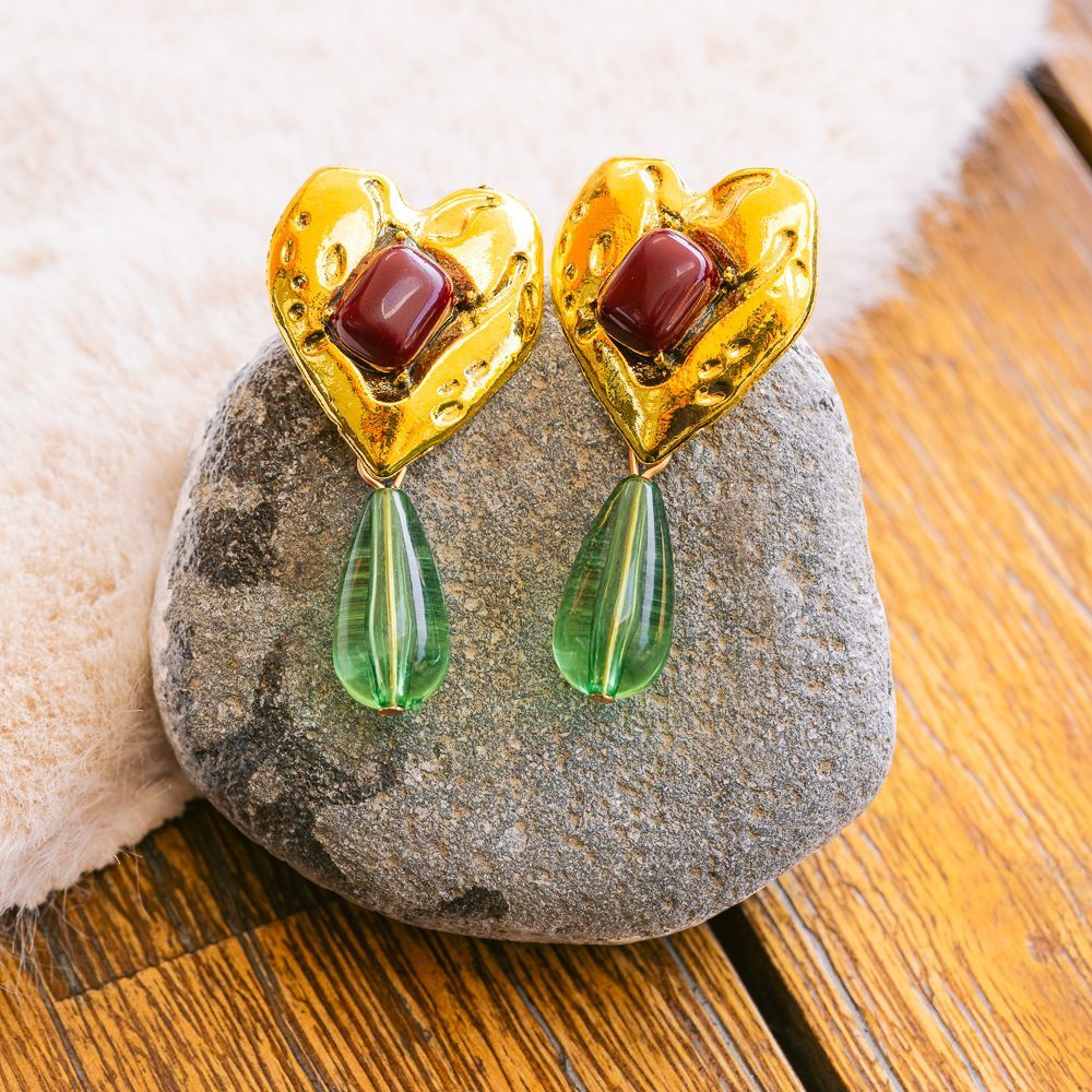 Pendientes de oro en forma de corazón con piedras rojas y lágrimas verdes que descansan sobre una piedra gris.