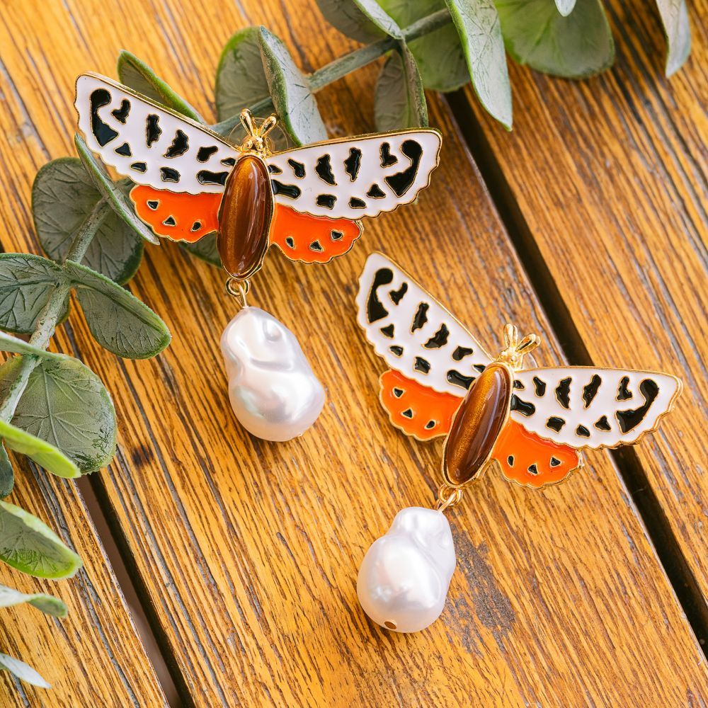 Pendientes con forma de mariposa con alas naranjas, negras y blancas sobre una superficie de madera con hojas verdes.