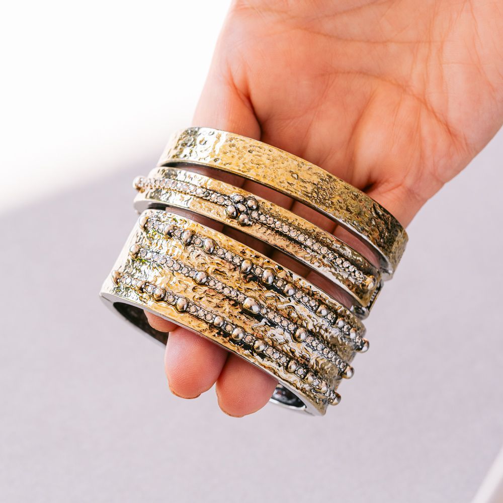 Una mano sostiene brazaletes apilados de oro y diamantes de imitación sobre un fondo claro.