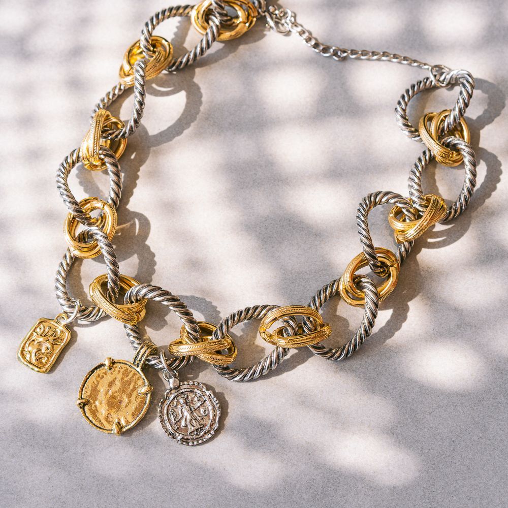 Collar de cadena de oro y plata con colgantes de monedas sobre un fondo estampado claro.