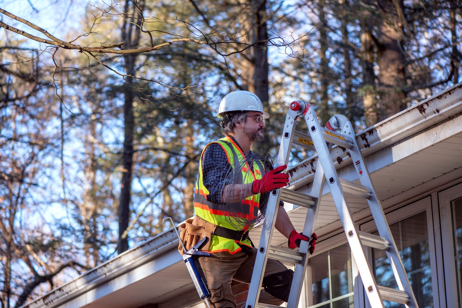 A man is standing on a ladder, wearing a safety helmet and vest, while performing gutter services.
