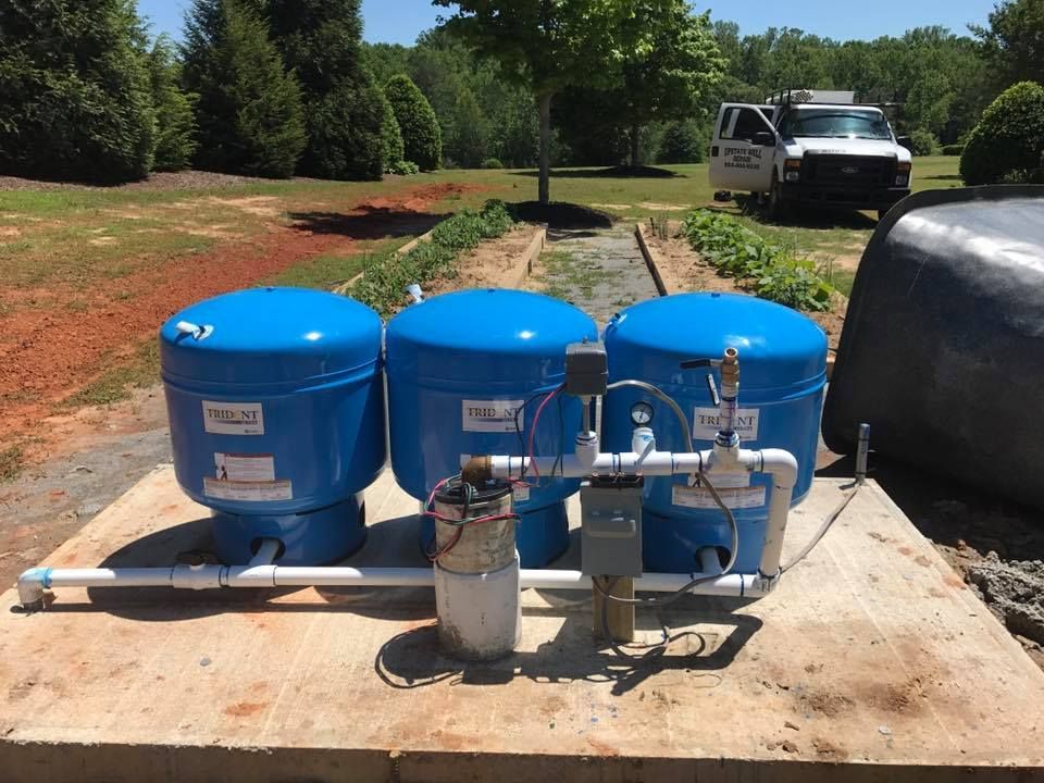 Three blue water tanks on a concrete pad with connected pipes, filter, and control box, outdoors near a truck.