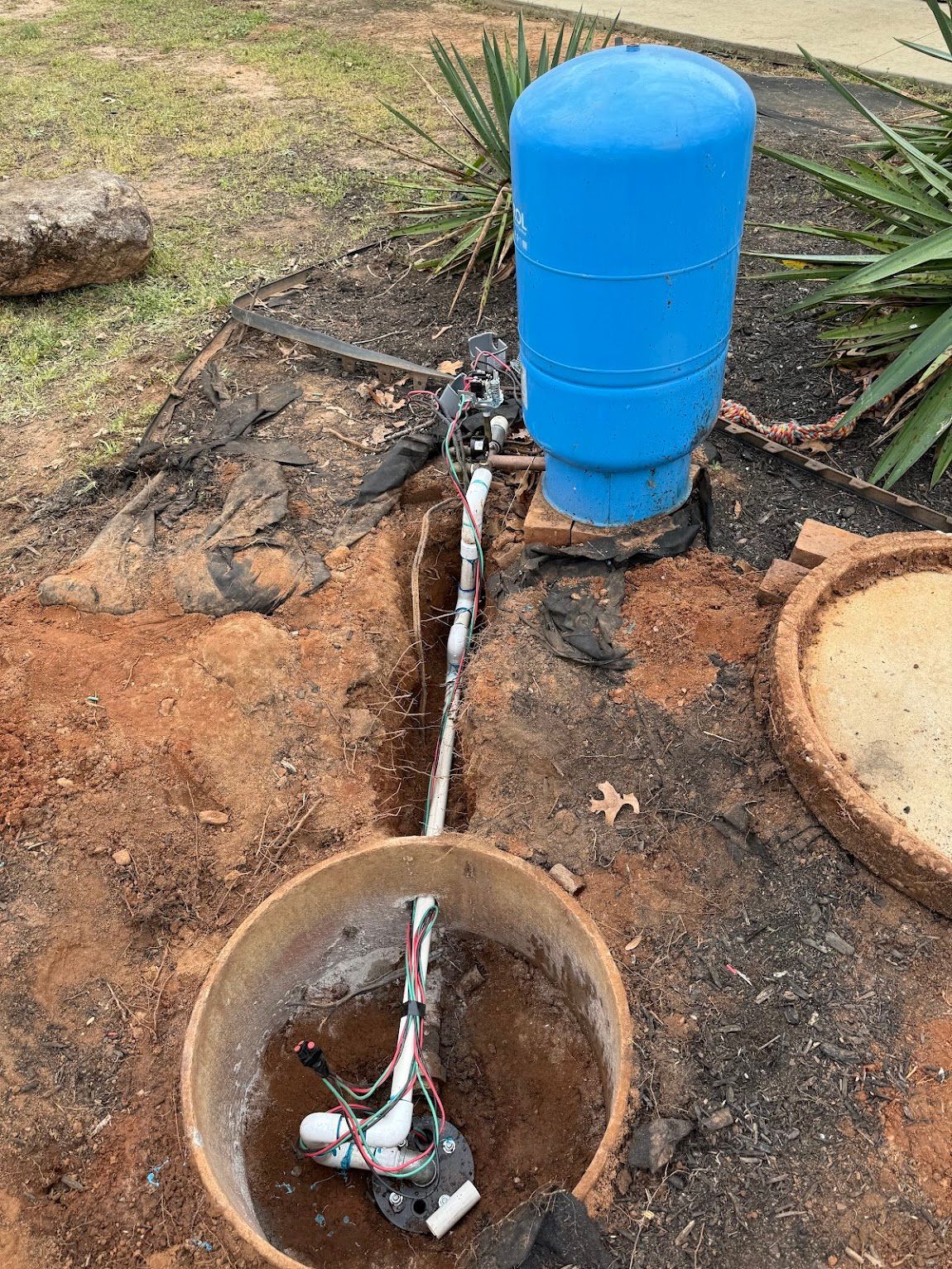 Well pump components: blue tank, exposed pipes, in-ground well and access lid, surrounded by dirt and landscaping.