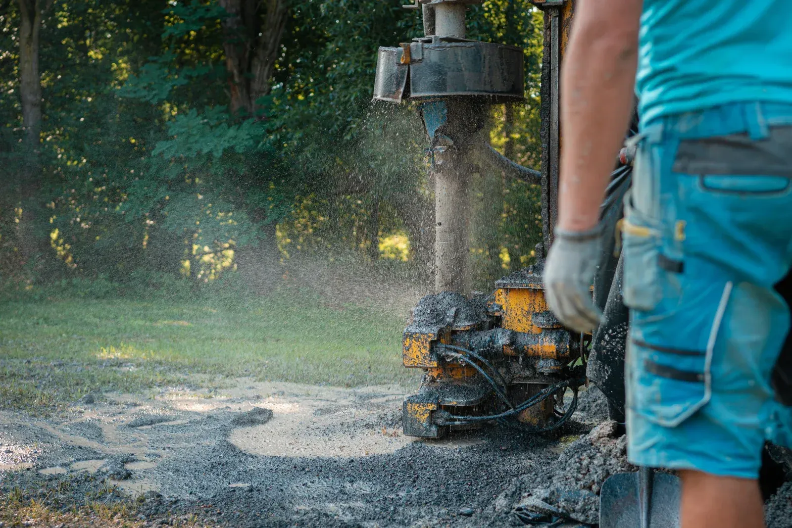 Man operating drilling rig, spraying water and mud. Green trees in background.