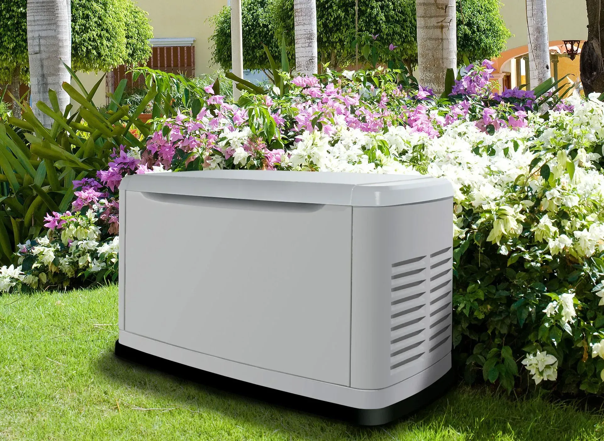 A gray home standby generator sits on a green lawn in front of a garden with purple and white flowers.