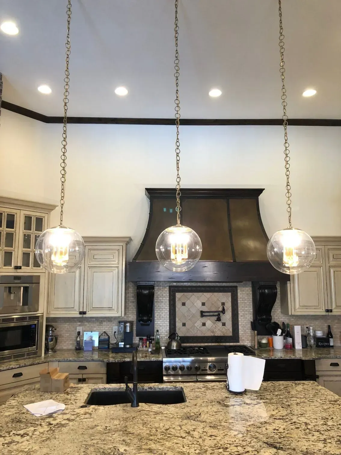 A kitchen island with three glass globe pendant lights hanging over a granite countertop, in front of a stove and vent.