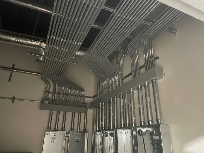 A series of metal electrical conduits run along a wall and ceiling, connecting to junction boxes in an industrial setting.