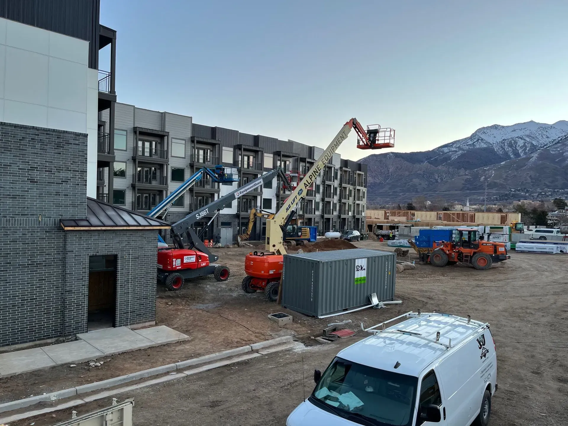 A construction site with several cherry pickers, a shipping container, and heavy machinery, set against a mountain backdrop.