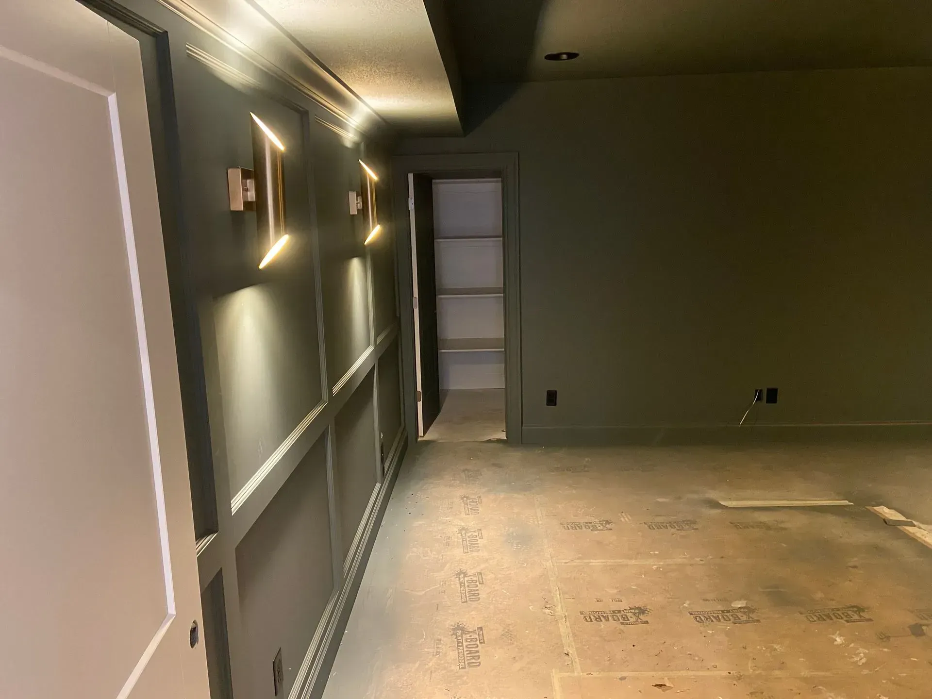 A dimly lit room with gray paneled walls, two brass wall sconces, a small doorway to a pantry, and an unfinished floor.