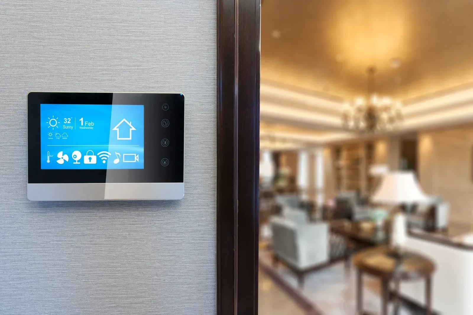 A smart home control panel mounted on a textured gray wall, next to a doorway overlooking a blurred, elegant living room.