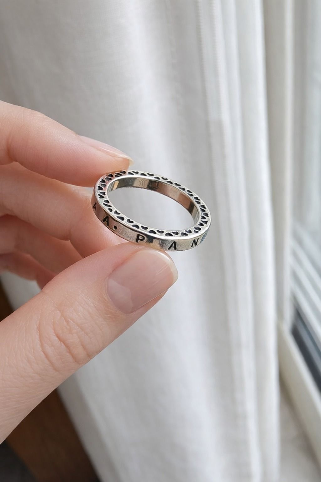 Una mano con un anillo de plata con pequeñas piedras junto a una ventana y una cortina blanca.