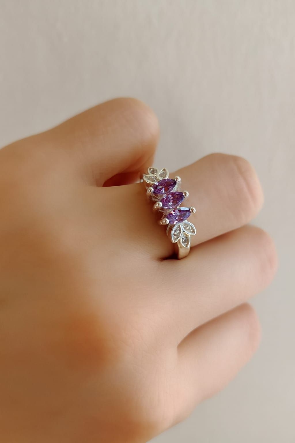 Mano que lleva un anillo de plata con piedras preciosas moradas y transparentes sobre un fondo beige suave.