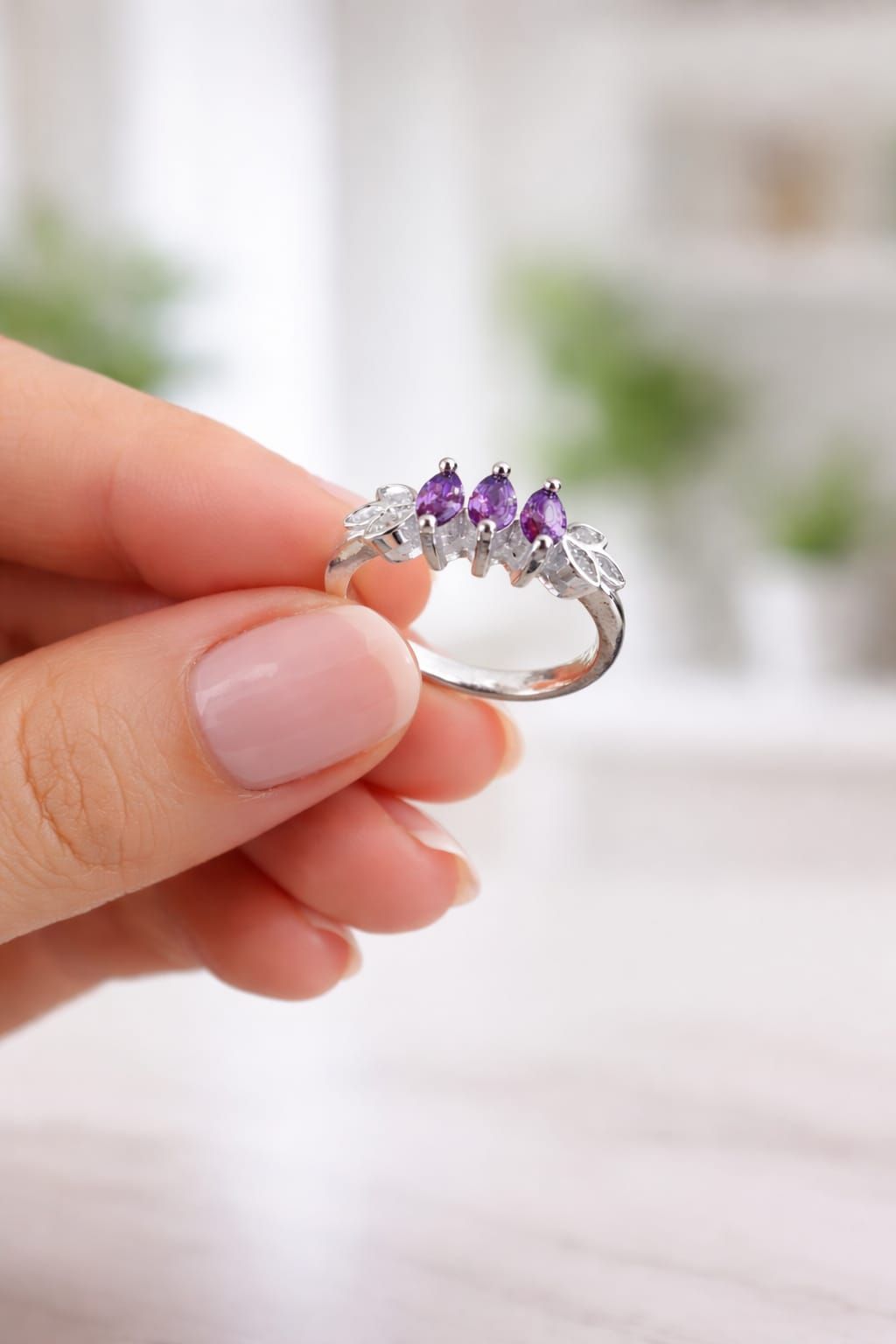 Mano que lleva un anillo de plata con piedras preciosas moradas y transparentes sobre un fondo beige suave.