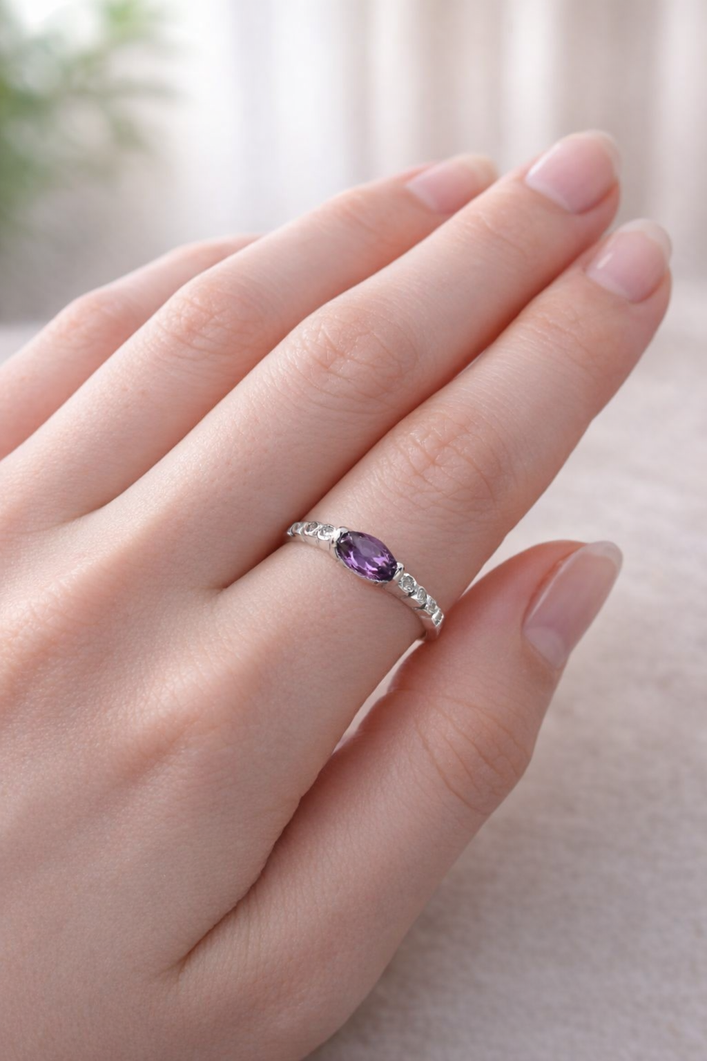 Mano con un anillo de plata con una gema púrpura sobre un fondo beige suave.