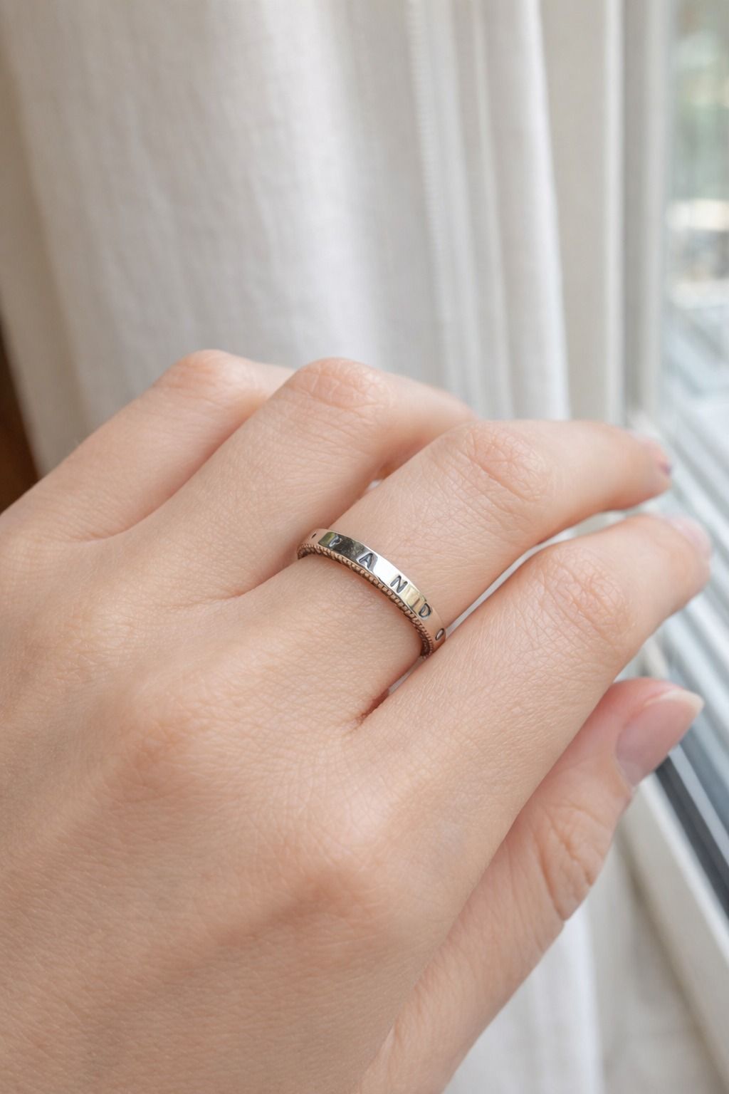 Una mano con un anillo de plata con pequeñas piedras junto a una ventana y una cortina blanca.