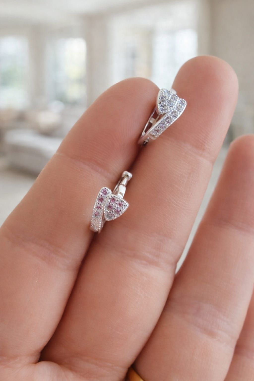 Dos pendientes plateados en forma de corazón sostenidos entre los dedos, con un fondo interior desenfocado.