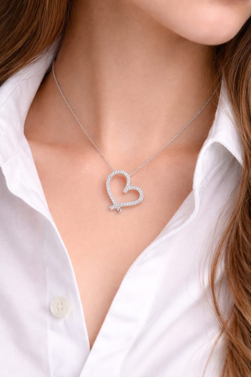 Mano sosteniendo un collar con colgante de corazón de plata con pequeñas piedras transparentes sobre un fondo de mármol.