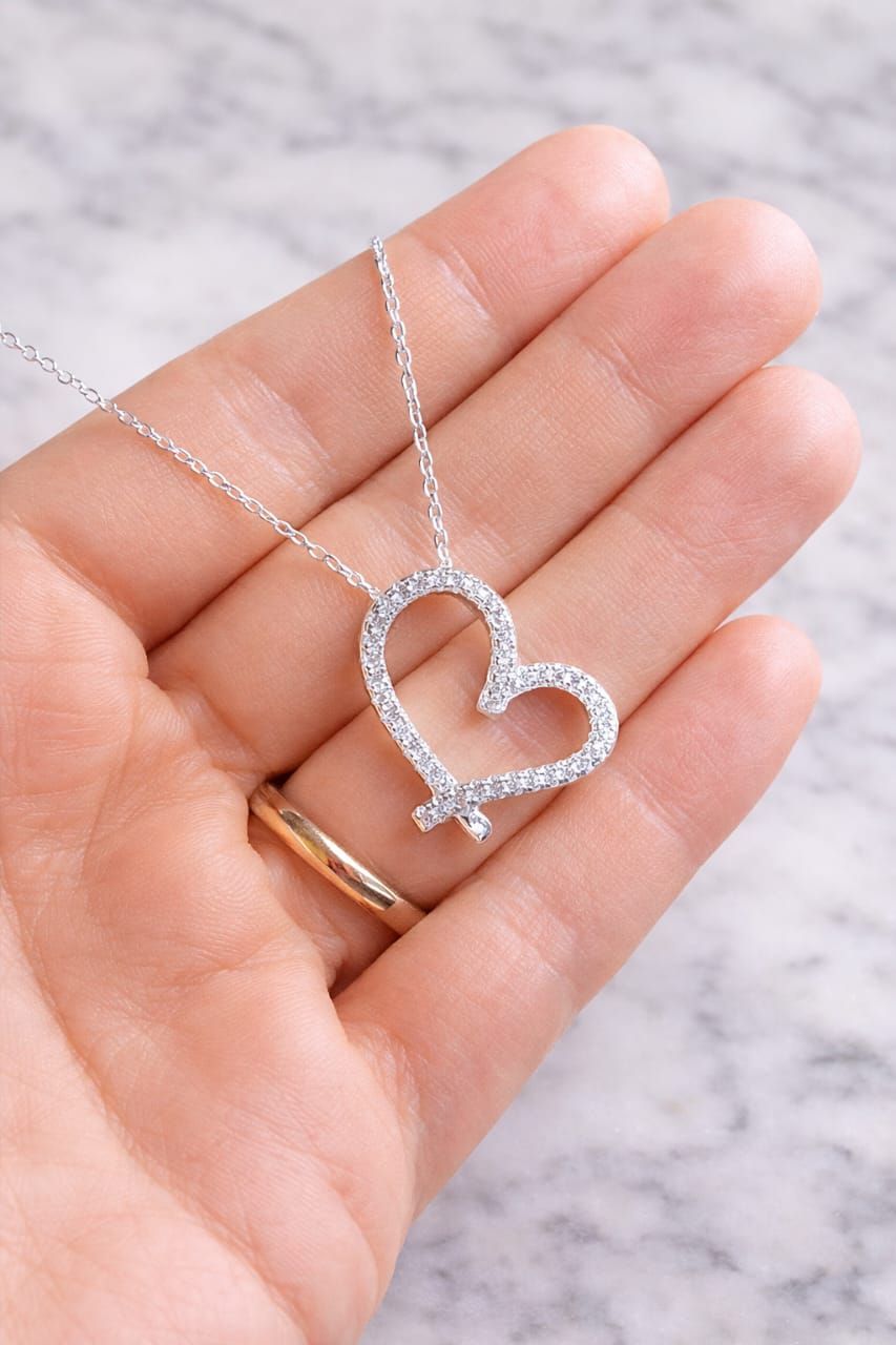 Mano sosteniendo un collar con colgante de corazón de plata con pequeñas piedras transparentes sobre un fondo de mármol.