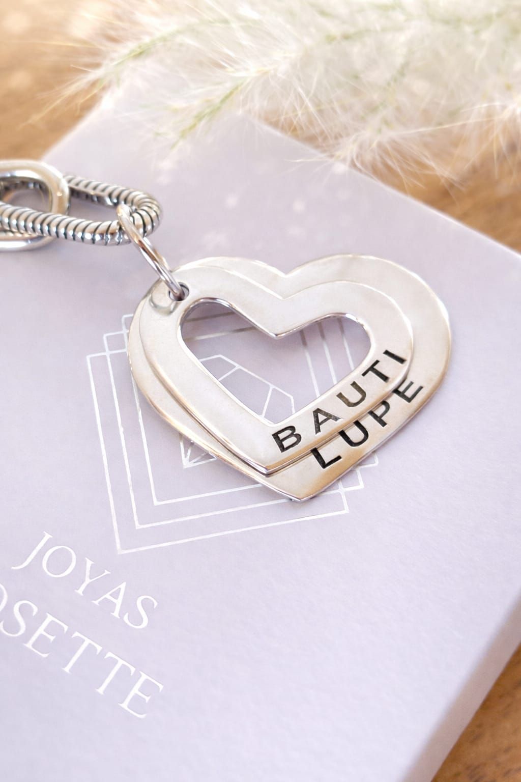 Collares de plata personalizados en una mano, con colgantes en forma de corazón y los nombres “Ciro” y “Valentina”.
