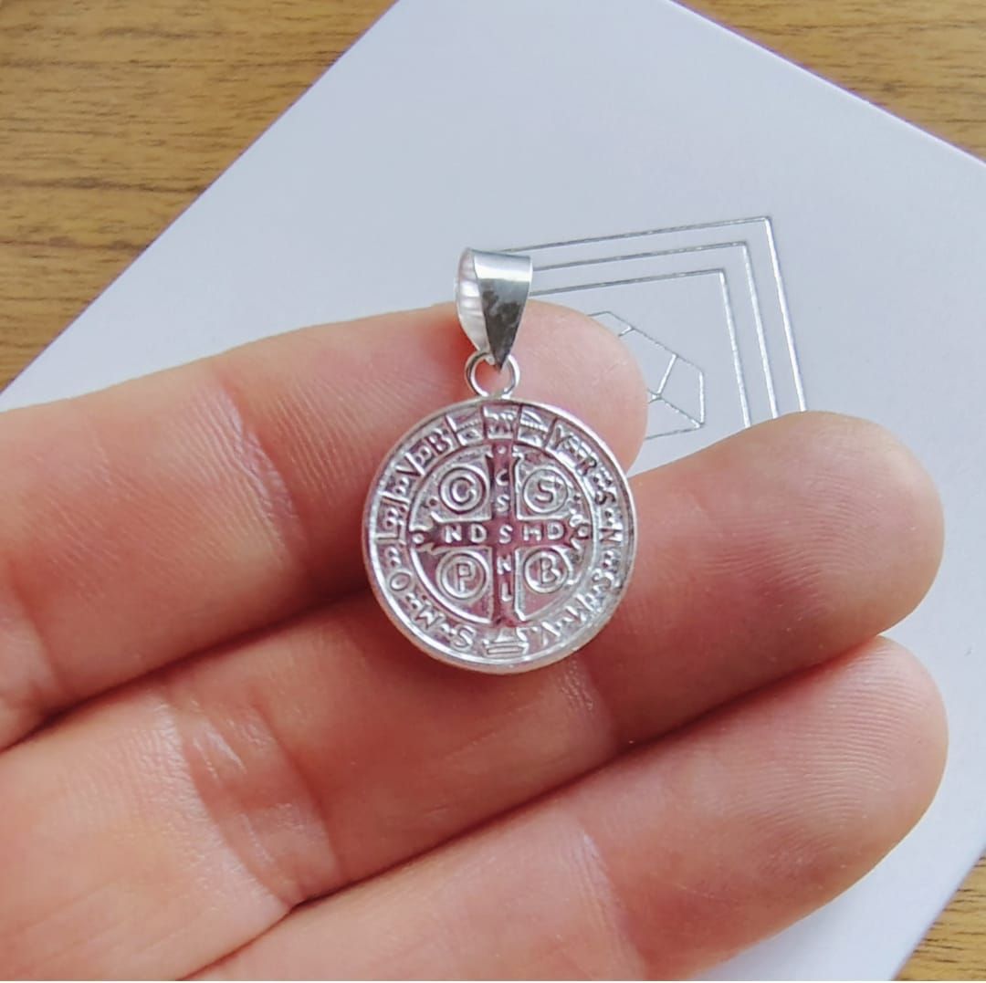 Pequeño colgante redondo de plata con un símbolo grabado, sostenido entre los dedos sobre un fondo de madera.