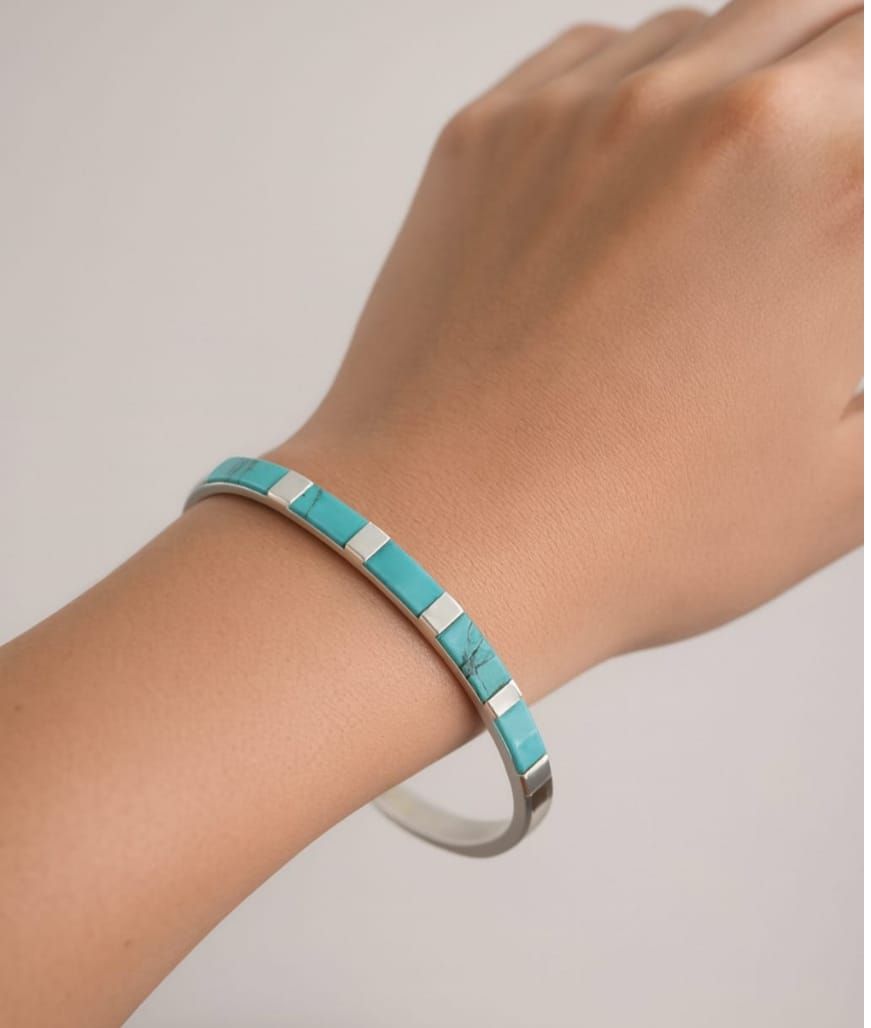 Una mano con una pulsera turquesa y plateada sobre un fondo neutro.