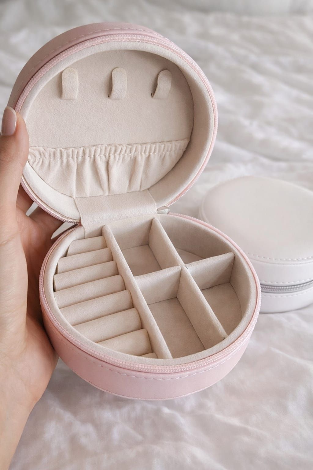 Caja de joyería redonda de color rosa abierta sobre una cama, que deja ver compartimentos color crema.