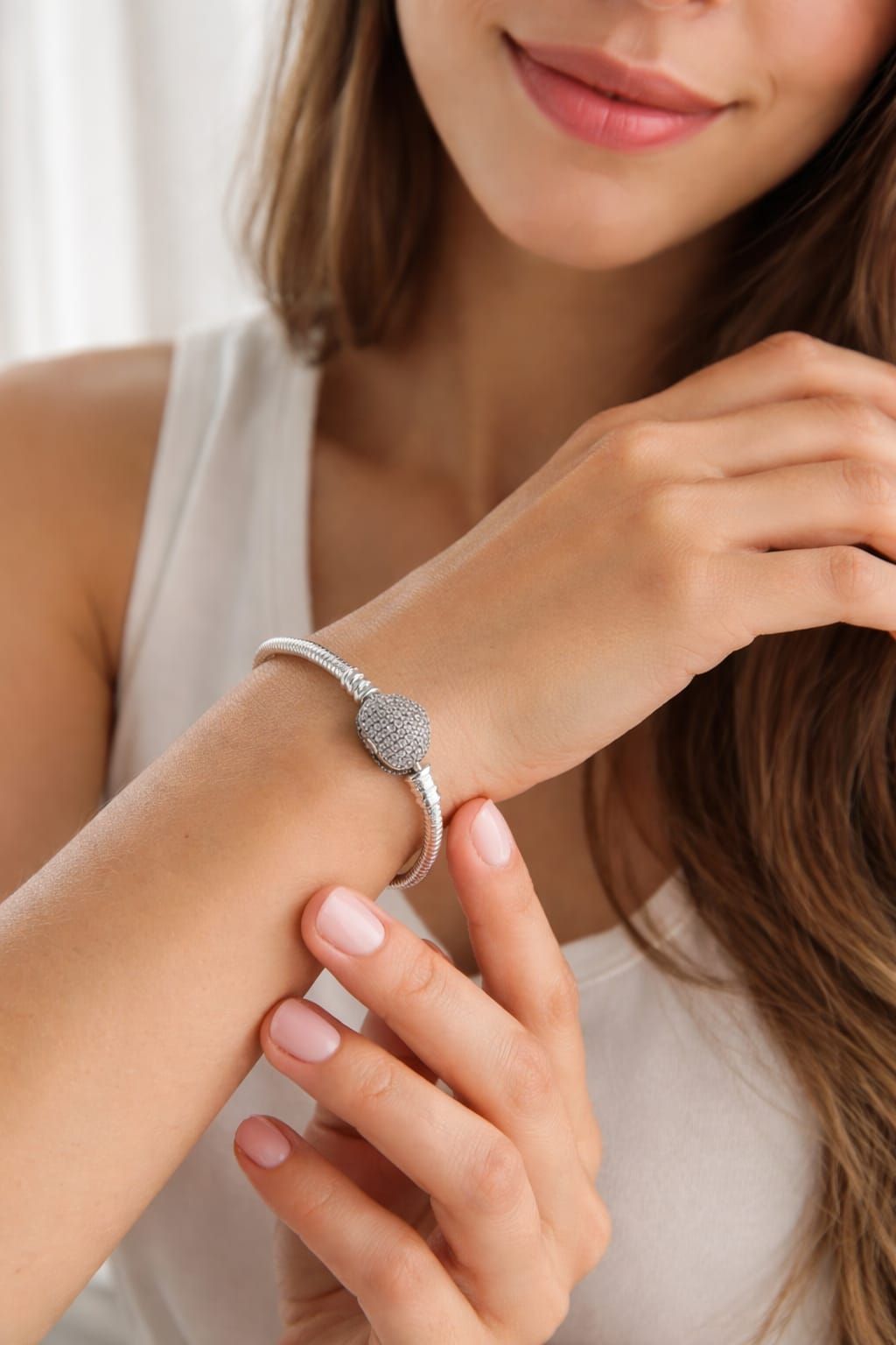 Mujer abrochándose una pulsera de plata en la muñeca, vestida con una blusa blanca sin mangas.