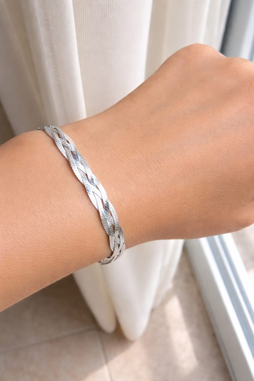 Pulsera trenzada de plata en una muñeca junto a una cortina blanca y una ventana.