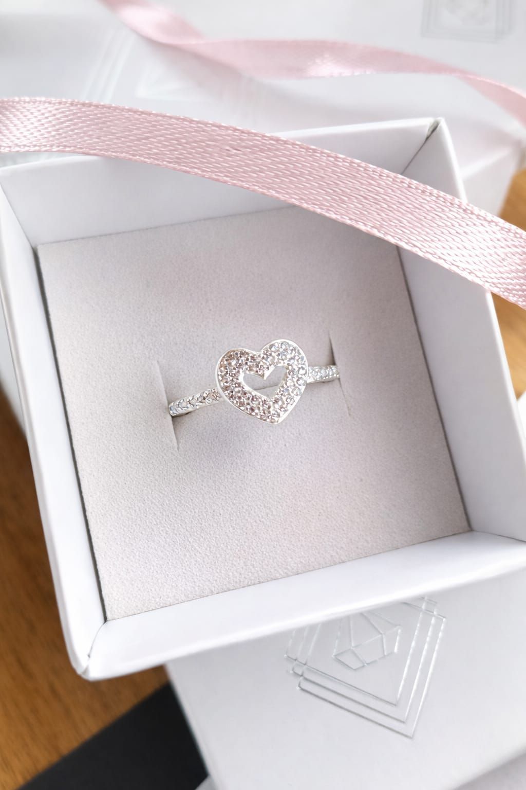 Mano con un anillo plateado en forma de corazón con pequeñas piedras, sobre un fondo interior suavemente desenfocado.