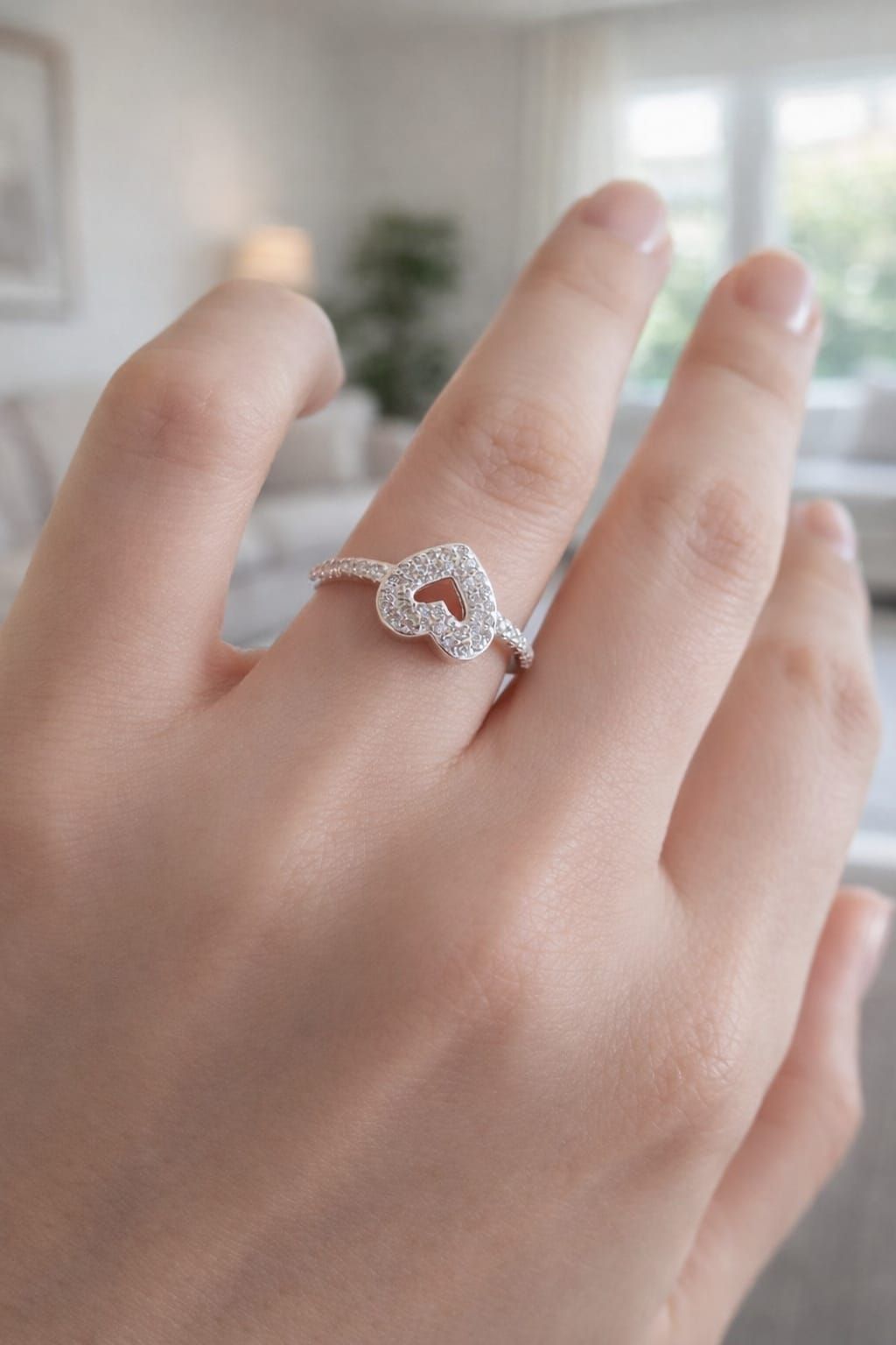 Mano con un anillo plateado en forma de corazón con pequeñas piedras, sobre un fondo interior suavemente desenfocado.