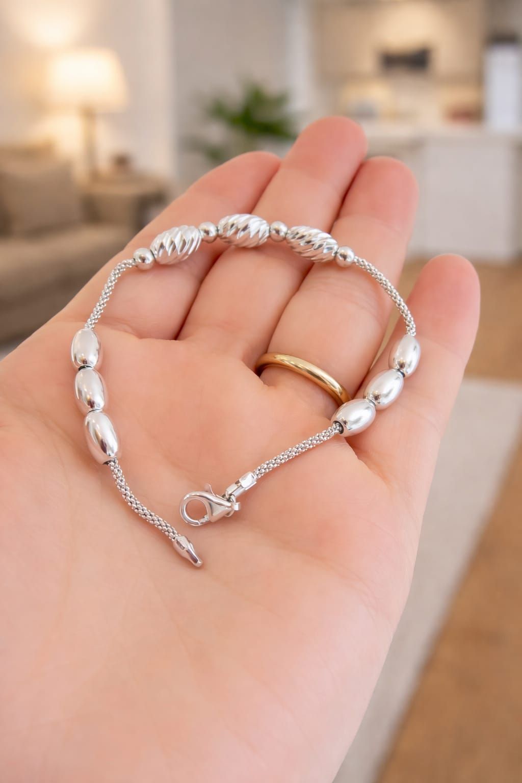 Pulsera de cuentas plateadas sobre una mano, con un anillo dorado en primer plano y un fondo de habitación suavemente