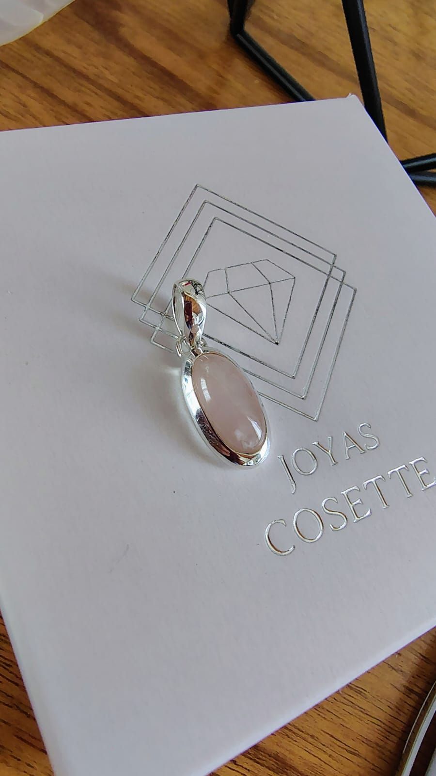 Una mano sostiene un pequeño colgante de plata con una piedra ovalada de color rosa pálido cerca de una ventana luminosa.